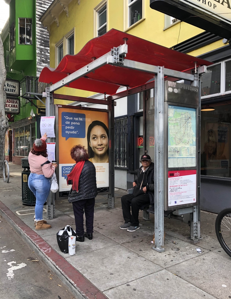 A woman shows signs about cancelled bus routes to another woman who had been sitting at a bus stop at 24th and Folsom streets waiting for the 48 Quintara/24th St. bus on April 8.