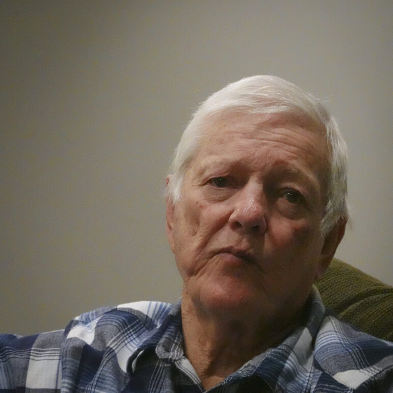 close-up of a white-haired man wearing a plaid shirt looking pensive and facing the camera