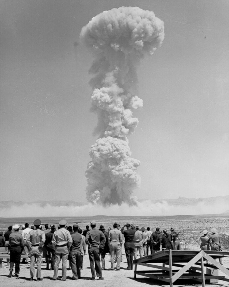 black-and-white photo of a crowd of people observing a large mushroom cloud in the desert 