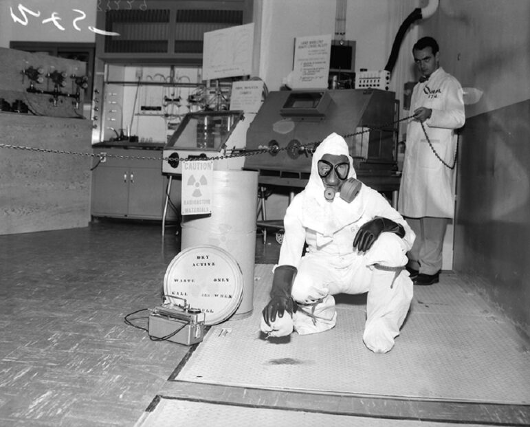 two men in a laboratory, one in a white lab coat and another in a gas mask and full-body hazard suit, posing in a black-and-white photo