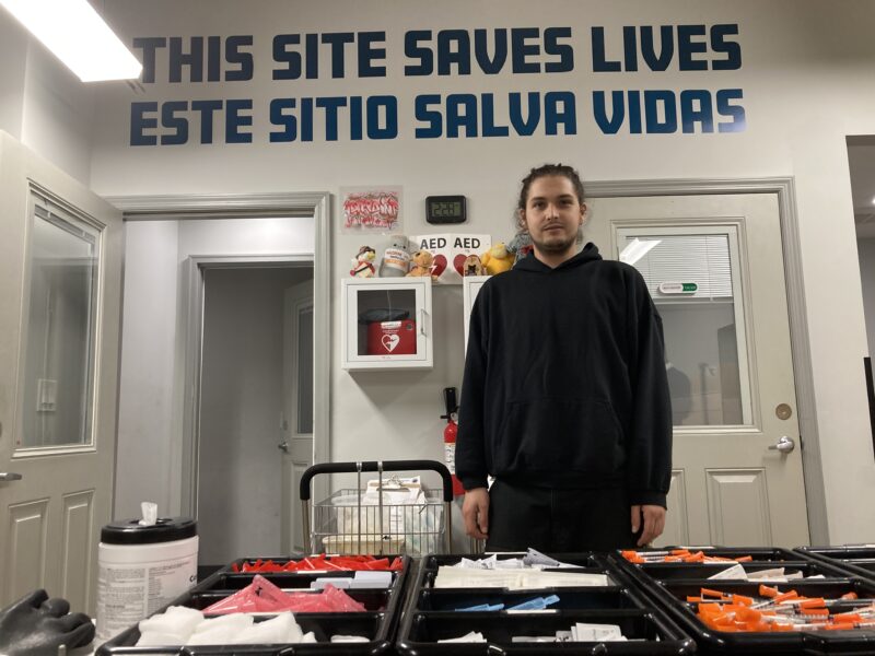 A man wearing dark clothing stands behind a table with syringes and trays of other medical supplies. The words "THIS SITE SAVES LIVES" above "ESTE SITIO SALVA VIDAS" are painted in blue in large letters high on the wall behind him.