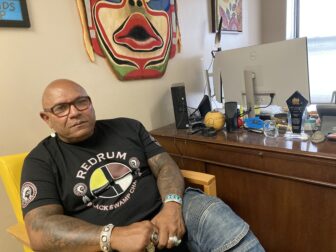 A man wearing a dark T-shirt and jeans sitsin a yellow chair next to a wooden desk in an office with colorful artwork on the wall behind him.