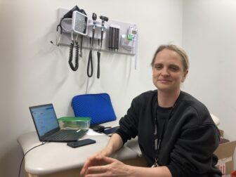 A woman with blonde hair pulled back and wearing a dark pullover sits at a desk with an open laptop and medical equipment hanging on the wall beside her.