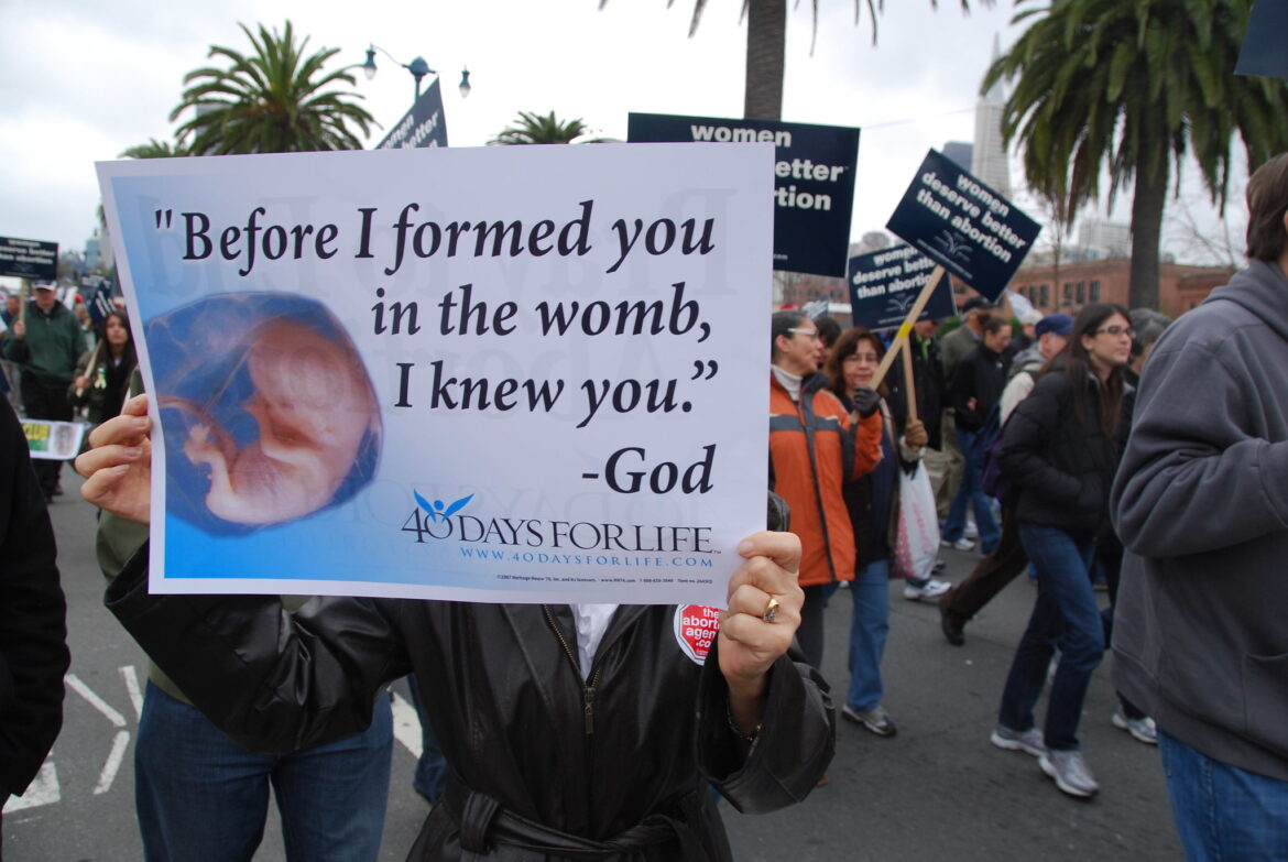 A political demonstrator holds up a sign that reads, “Before I formed you in the womb, I knew you. -God”