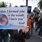 A political demonstrator holds up a sign that reads, “Before I formed you in the womb, I knew you. -God”