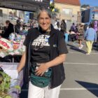 A woman wearing a dark top and white pants stands next to a table displayng informational pages at a community festival.