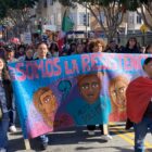 Throngs of people carrying signs and flags walk down an urban street behind four people holding a large blue banner reading "Somos La Resistencia" in Spanish in large pink letters, with a smaller English translation reading "We are the resistance."