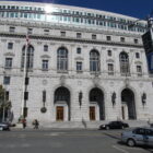 A large gray masonry building features arched windows and doorways. Light reflects off the glass of a modern building behind it.