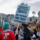 In a crowd, a person with teal hair in a red shirt holds up a poster that says "IMMIGRANT AND WORKERS RIGHTS ONE STRUGGLE ONE FIGHT"