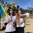 Two young women facing forward wear bright colored headdresses of artificial flowers and white blouses with red and burgundy embroidery. They are standing in a park and one carries a yellow and bright blue flag.